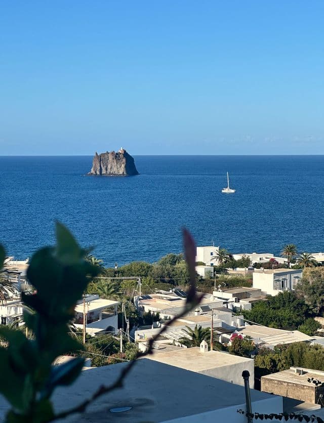 Un isolotto roccioso e una barca a vela solitaria sul mare blu intenso, visti dall'alto di un villaggio costiero con edifici bianchi.