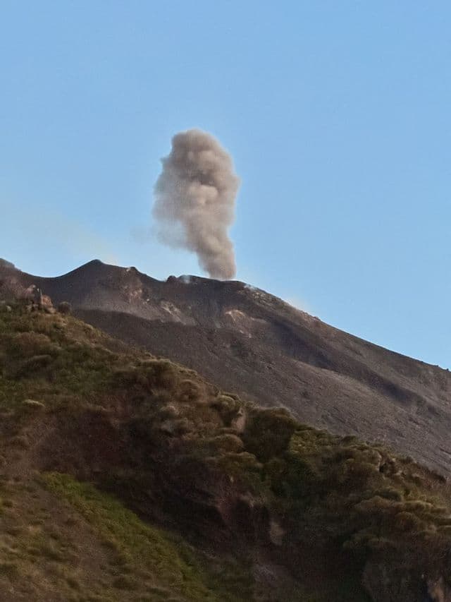 Eine graue Rauchsäule steigt aus dem Krater eines dunklen Vulkans unter einem klaren blauen Himmel auf, mit einem grünen Hang im Vordergrund.