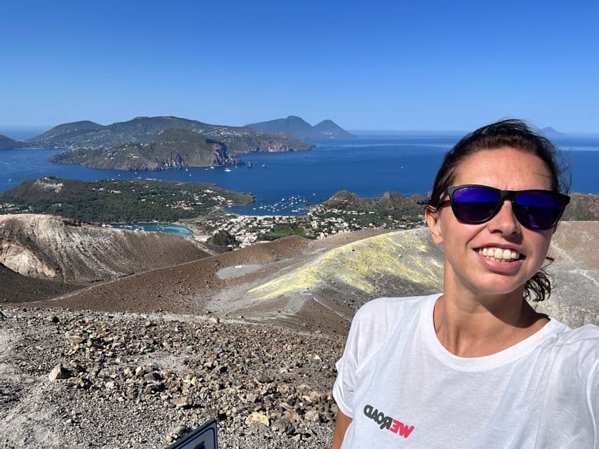 Una donna sorridente che indossa occhiali da sole si scatta un selfie da un cratere vulcanico con vista su isole e una città costiera sul mare.
