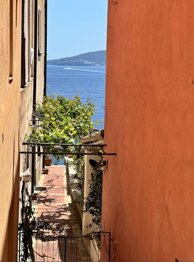 Una vista lungo un vicolo stretto e soleggiato tra edifici in terracotta, che si apre sul mare blu con una barca in lontananza.