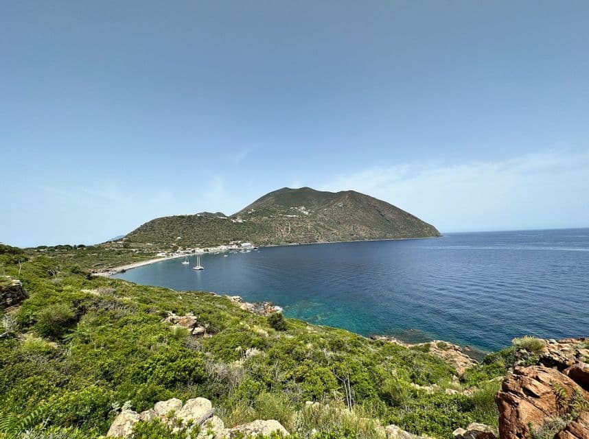 Vista dall'alto di una baia costiera con barche a vela, un piccolo villaggio e una grande collina verde sotto un cielo azzurro e limpido.