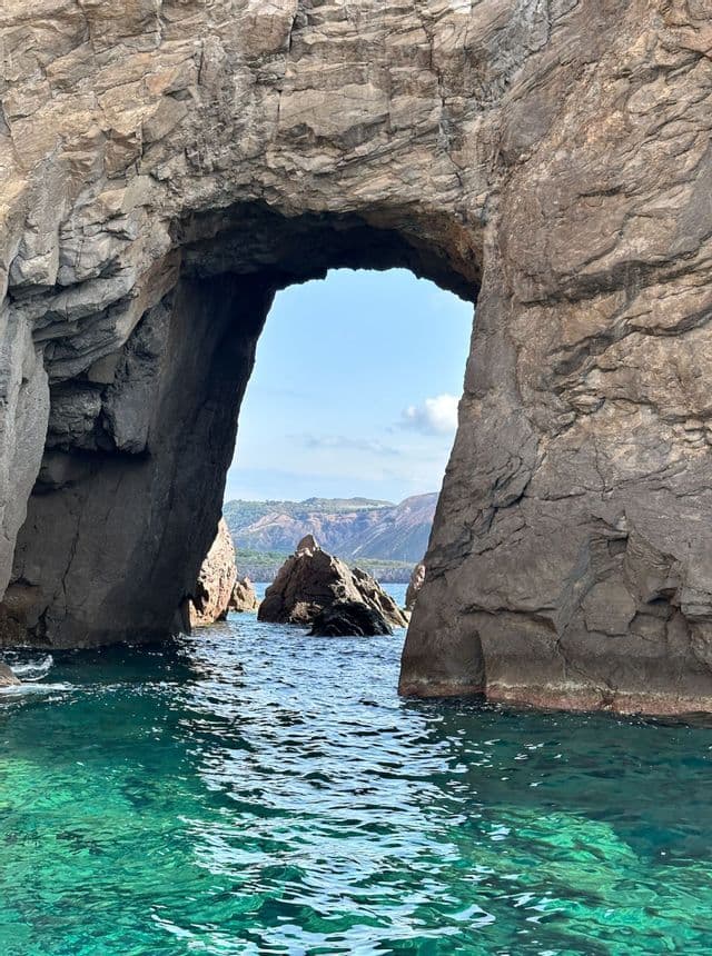 Vista dall'acqua, attraverso un grande arco di roccia naturale, verso la costa lontana sotto un cielo azzurro.