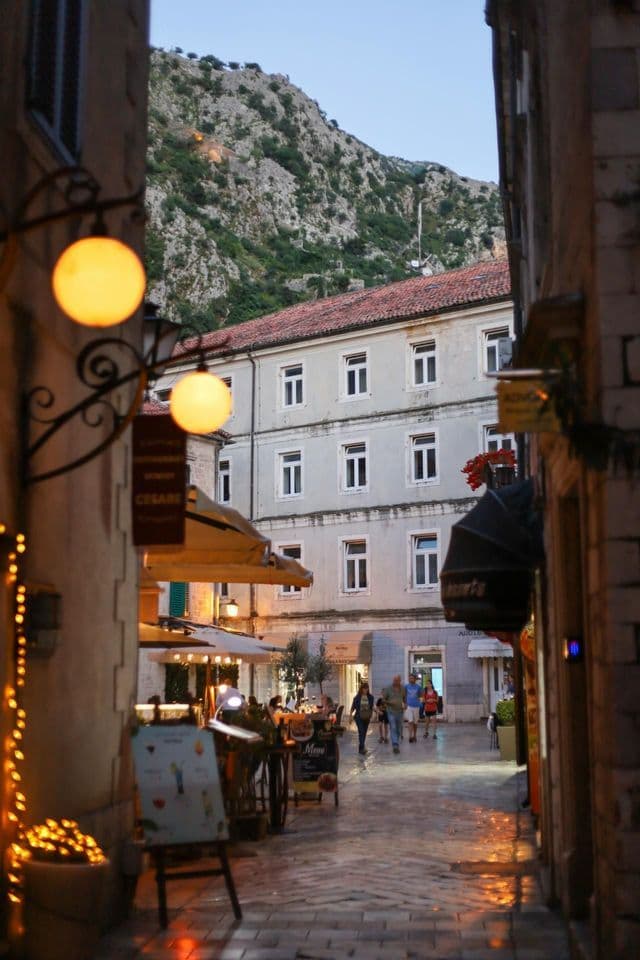 Eine schmale Kopfsteinpflasterstraße in der Dämmerung, erleuchtet von warmen Lichtern aus Cafés, mit flanierenden Menschen und einem Berg im Hintergrund.