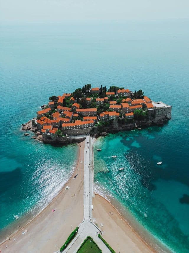 Luftaufnahme einer kleinen Insel mit rot gedeckten Gebäuden, die über einen Damm mit einem Sandstrand verbunden ist, umgeben von türkisfarbenem Wasser.