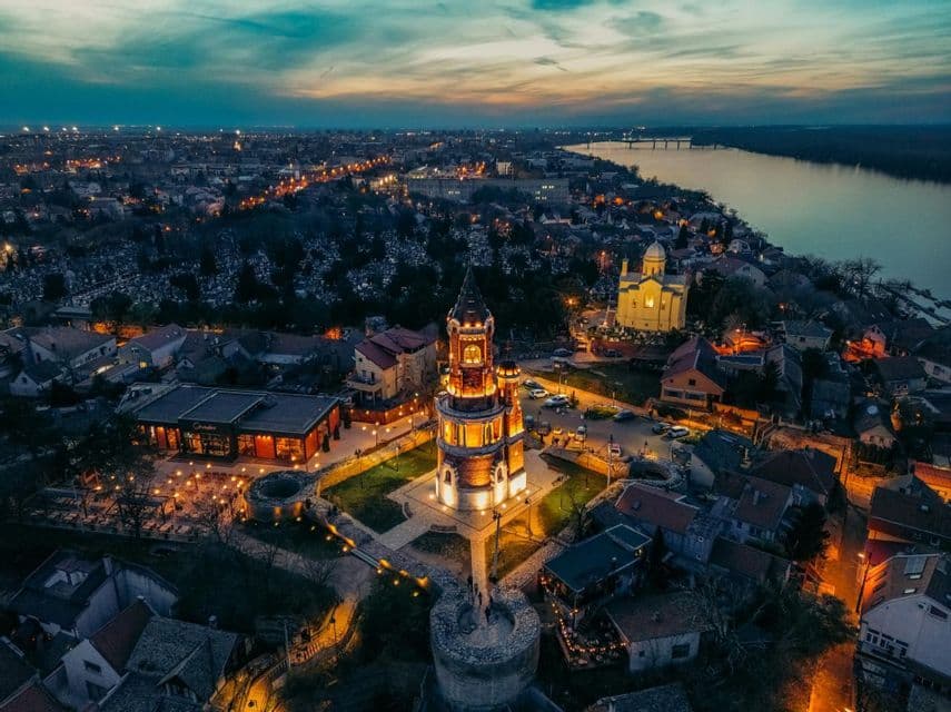 Blick aus der Luft auf einen beleuchteten historischen Turm in einer Stadt am Fluss bei Sonnenuntergang, mit in die Ferne reichenden Stadtlichtern.