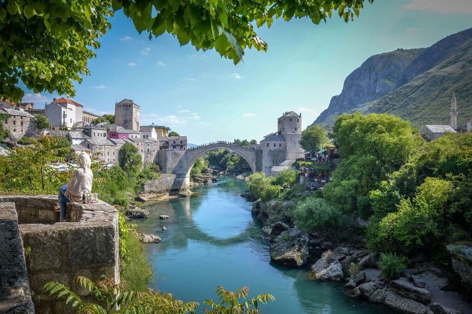 Eine blondhaarige Person sitzt auf einer Steinmauer und blickt auf eine historische Bogenbrücke, die einen türkisfarbenen Fluss in einer Altstadt überspannt.
