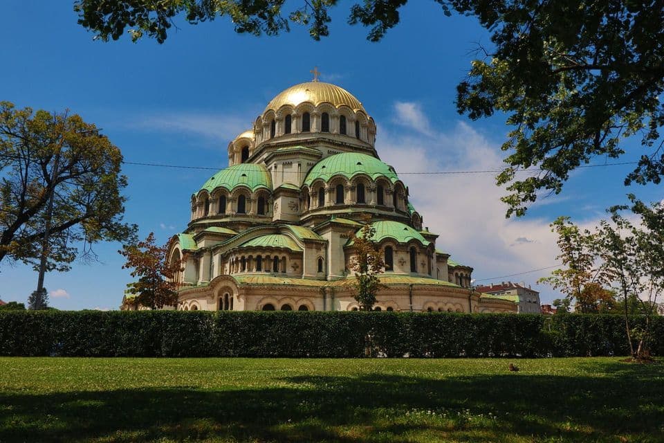 Eine große Kathedrale mit einer zentralen goldenen Kuppel und kleineren grünen Kuppeln, von einem Park hinter einer Hecke an einem sonnigen Tag gesehen.