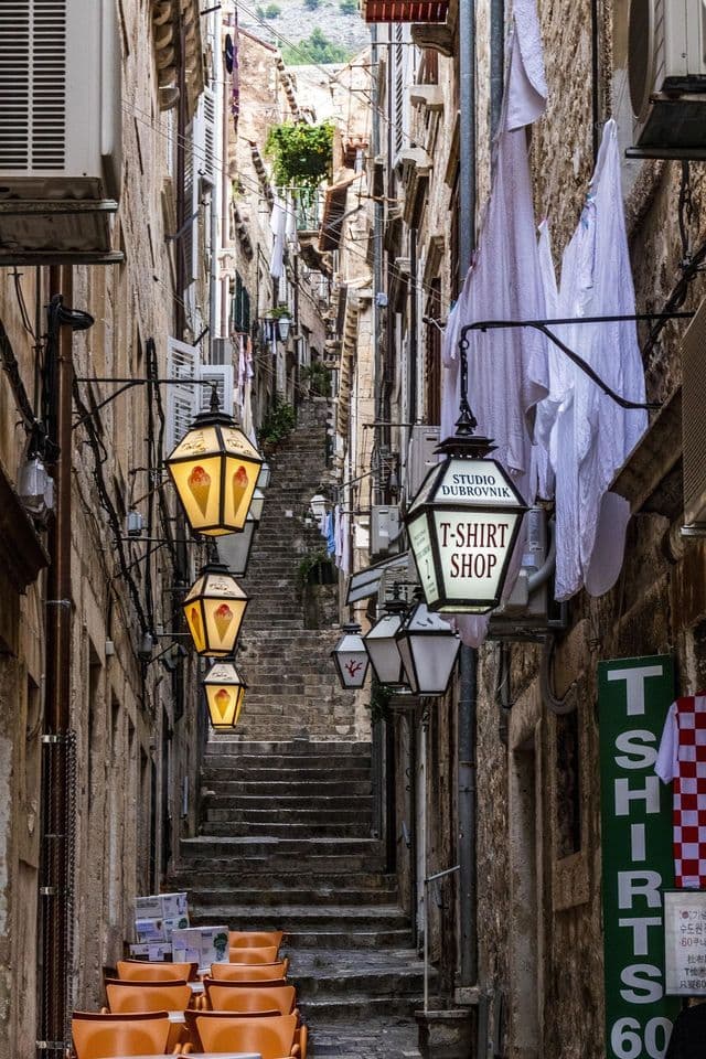 Eine steile Steintreppe windet sich durch eine schmale Gasse, gesäumt von Gebäuden, hängenden Laternen und trocknender Wäsche.