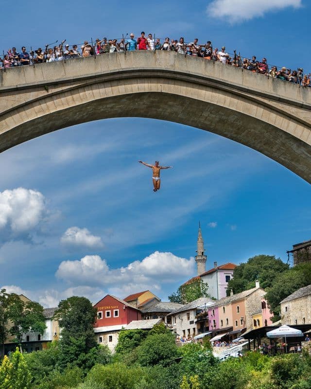 Ein Mann mit ausgestreckten Armen springt von einer hohen Steinbogenbrücke, während eine Menschenmenge von oben zuschaut und eine Stadt darunter liegt.