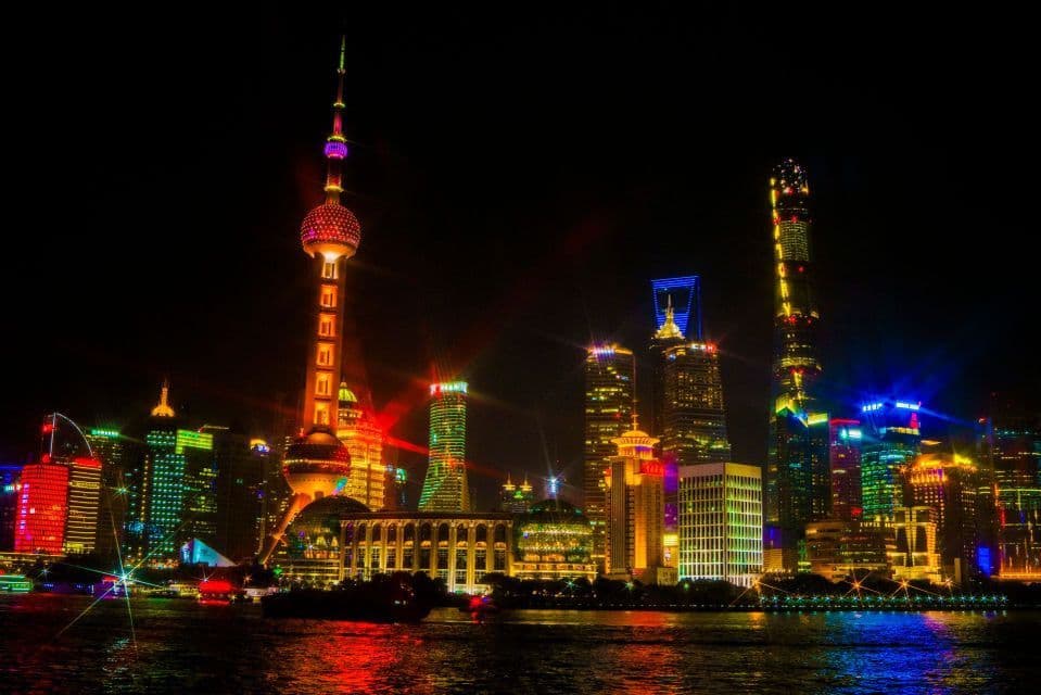 Uno skyline cittadino con un'alta torre e grattacieli illuminati da luci al neon colorate di notte, con riflessi sull'acqua sottostante.