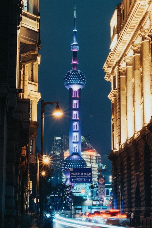 Una torre futuristica illuminata con luci blu e viola, incorniciata tra due edifici su una strada cittadina di notte.