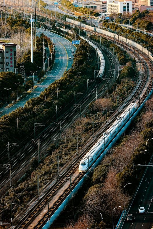 Vista aerea di un treno bianco ad alta velocità che viaggia su binari accanto a una strada tortuosa con auto e altre linee ferroviarie.