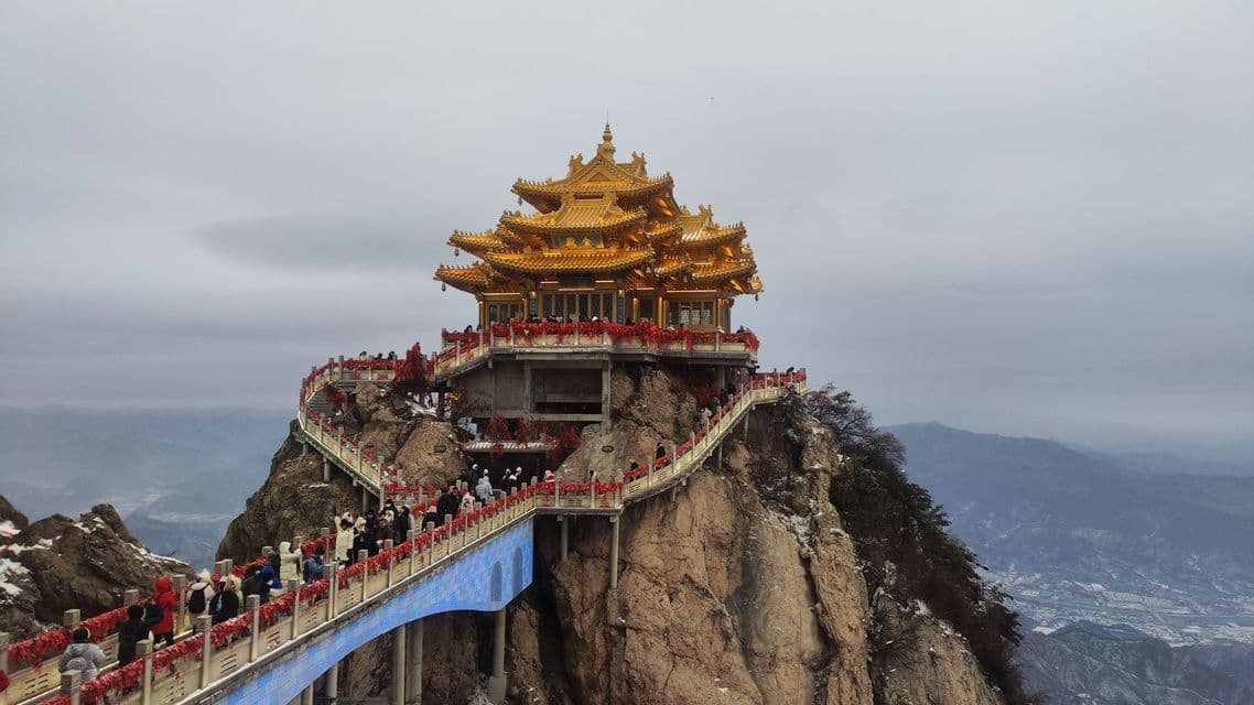 Un tempio d'oro si erge su una vetta montuosa rocciosa, con folle di persone su un ponte tortuoso che vi conduce sotto un cielo nuvoloso.