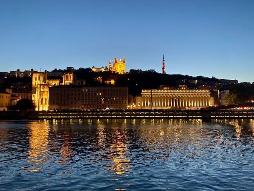 Uno skyline cittadino illuminato con una basilica su una collina, visibile oltre un fiume al crepuscolo con riflessi sull'acqua.