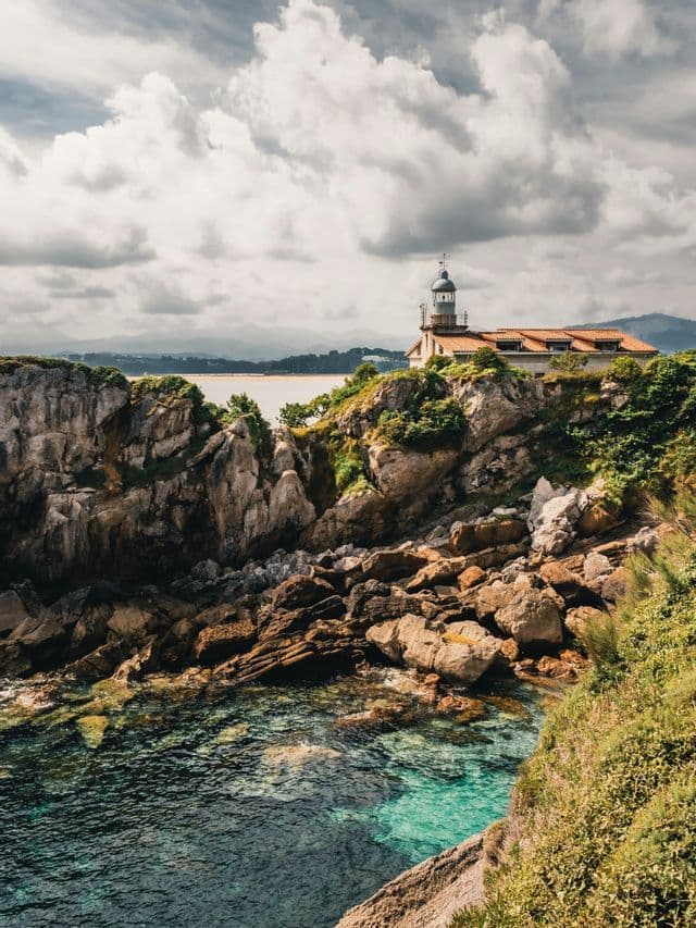 Un faro con techo de tejas se alza en un acantilado rocoso y vegetado, con vistas a aguas turquesas transparentes bajo un cielo nublado.