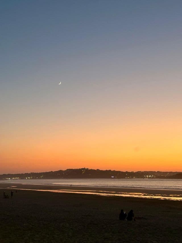 Una luna creciente cuelga en un cielo degradado de azul y naranja sobre una playa al anochecer, con personas silueteadas en la arena.
