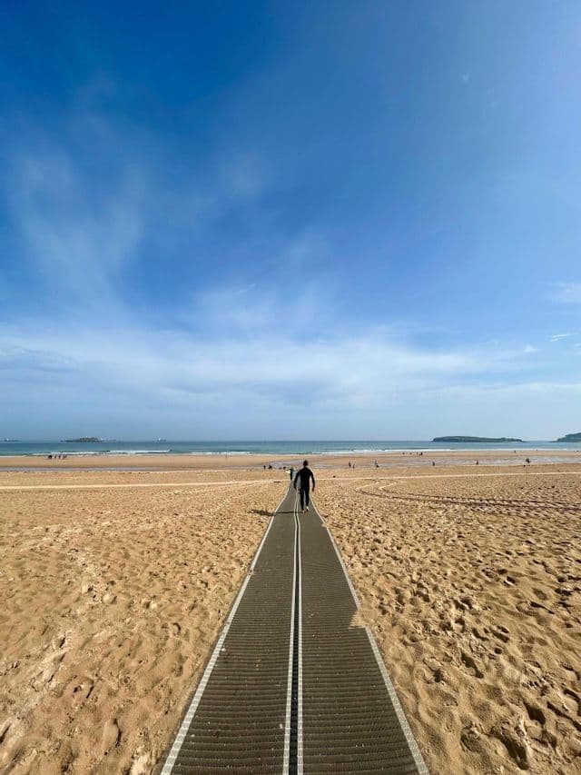 Una persona en traje de neopreno camina por un sendero a través de una playa de arena hacia el océano bajo un cielo azul.
