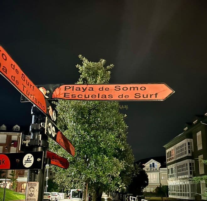 Un letrero de calle de noche con señales rojas que indican 'Playa de Somo' y 'Escuelas de Surf'.