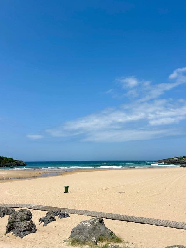 Un paseo marítimo de madera conduce a una amplia playa de arena junto al océano bajo un cielo azul parcialmente nublado.
