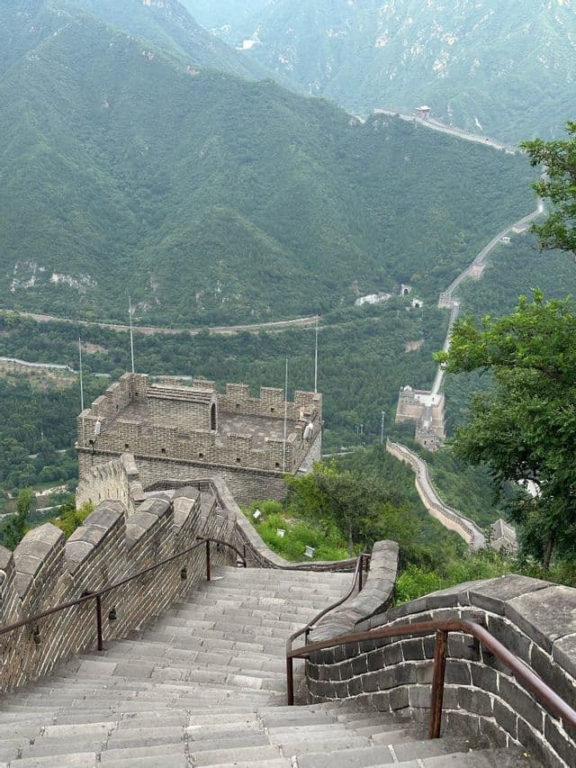 Vista dall'alto di una ripida scalinata in pietra su un lungo muro con una torre di guardia, che si snoda tra montagne verdi.
