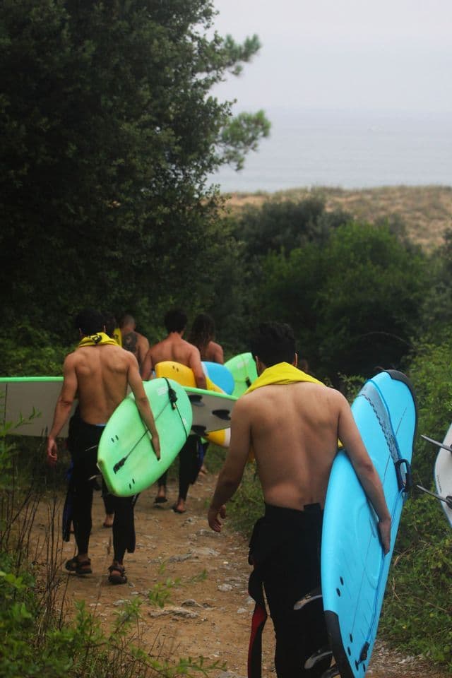 Un grupo de surfistas de WeRoad, en trajes de neopreno, caminando por un sendero con sus tablas hacia el océano.