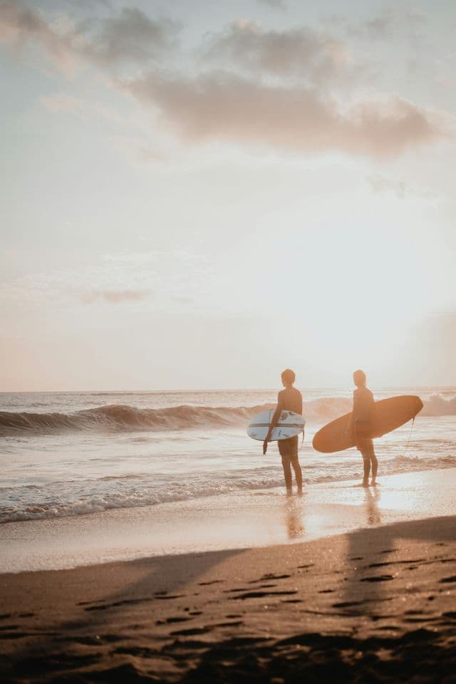 Dos surfistas con tablas de surf de pie a la orilla del agua en una playa de arena, observando las olas al atardecer.