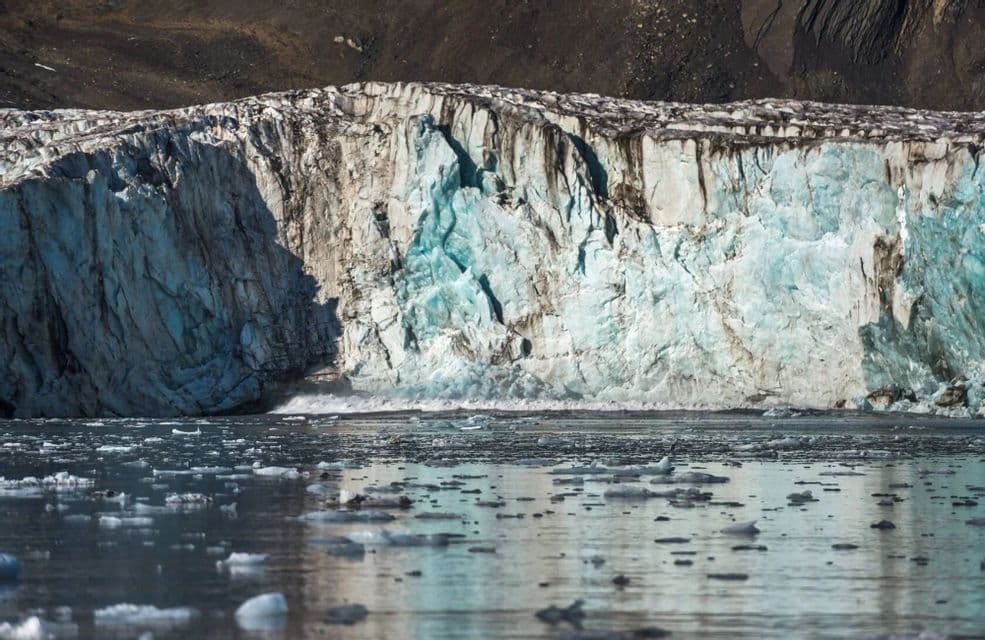 Una imponente pared de glaciar de hielo azul y blanco se eleva sobre un cuerpo de agua lleno de pequeños trozos de hielo flotantes.