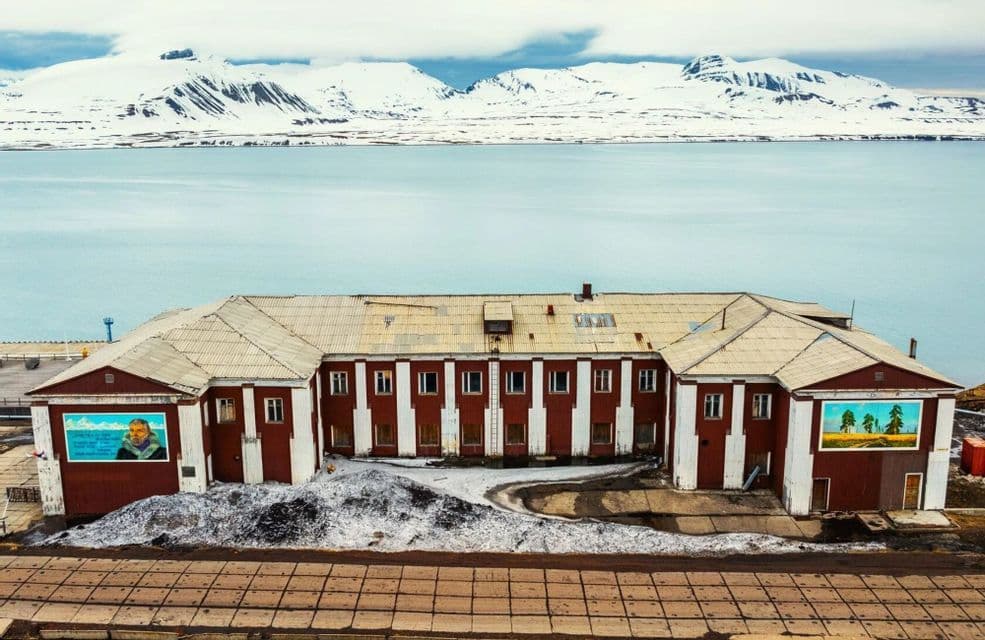 Un edificio rojo y blanco se encuentra en la orilla frente a una vasta extensión de agua y una cordillera nevada.