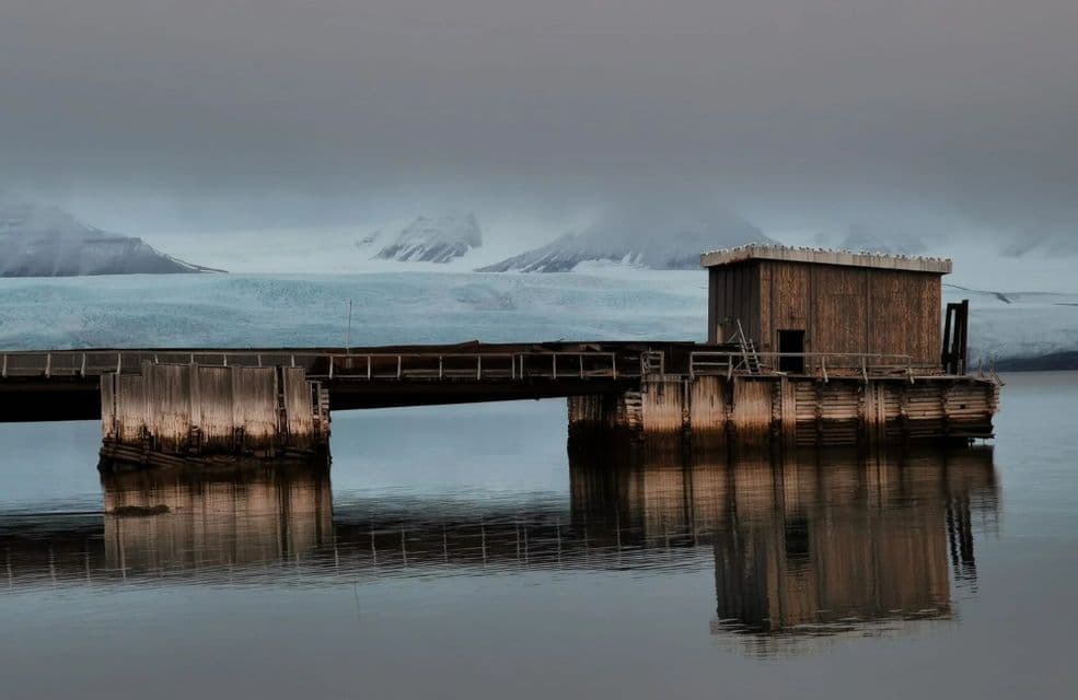 Un muelle de madera con una pequeña cabaña se refleja en aguas tranquilas frente a un vasto glaciar y montañas nevadas bajo un cielo nublado.