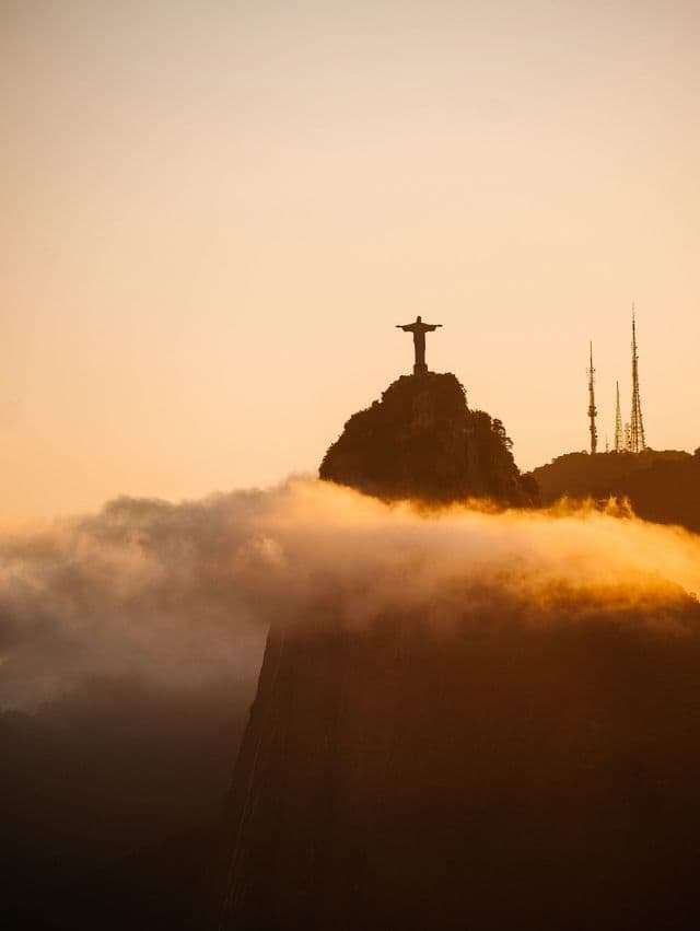 Una sagoma di una grande statua con le braccia aperte su una cima di montagna, che si erge sopra le nuvole contro un cielo dorato.