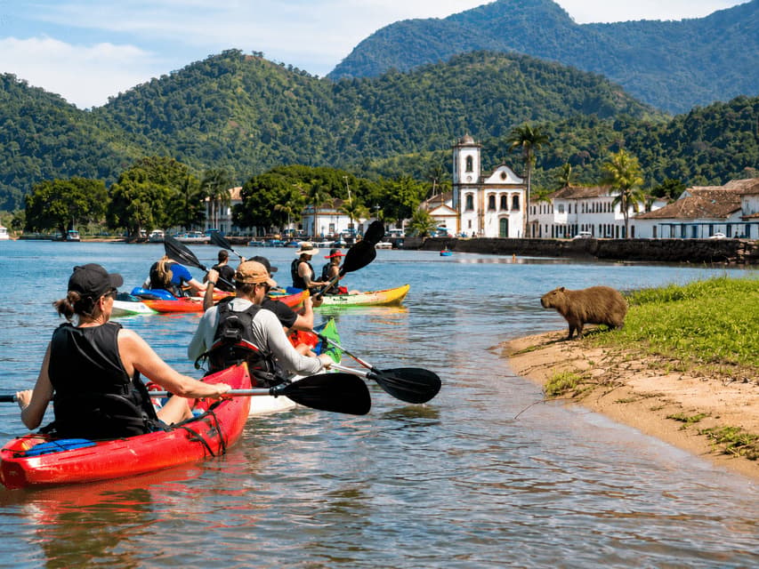 Un gruppo WeRoad in kayak su un fiume, osservando un capibara sulla riva con una città coloniale e montagne verdi sullo sfondo.
