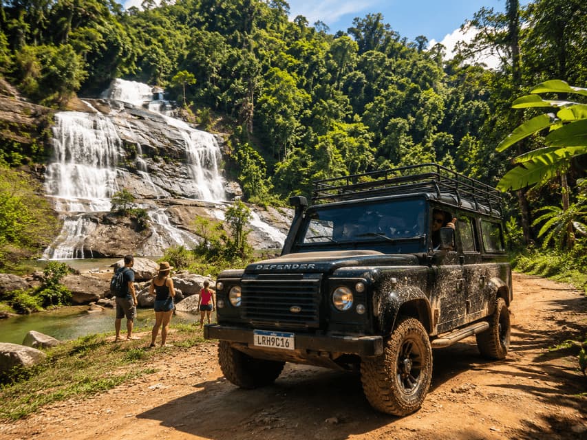 Un veicolo fuoristrada infangato è parcheggiato su un sentiero nella giungla, mentre un gruppo WeRoad si trova accanto a una grande cascata sullo sfondo.
