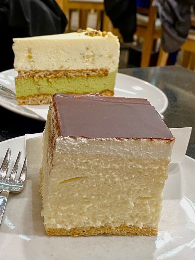 Un pastel de mousse cuadrado con cobertura de chocolate y un pastel de pistacho en capas servido en platos blancos.