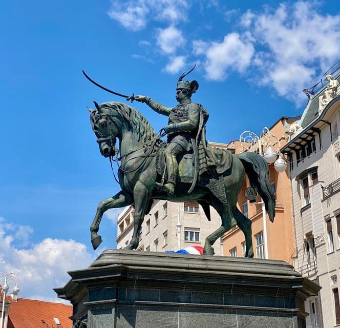 Una estatua ecuestre de bronce de un hombre uniformado levantando una espada, con un cielo azul y edificios de la ciudad de fondo.