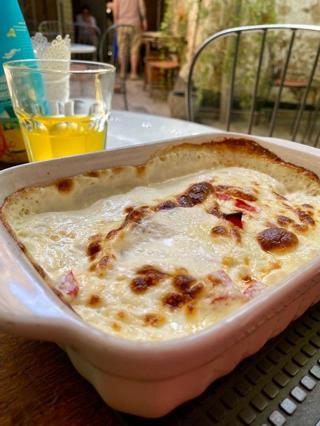 Primer plano de una cazuela horneada con cobertura de queso dorada en un plato blanco, junto a un vaso de zumo de naranja en una mesa de patio.