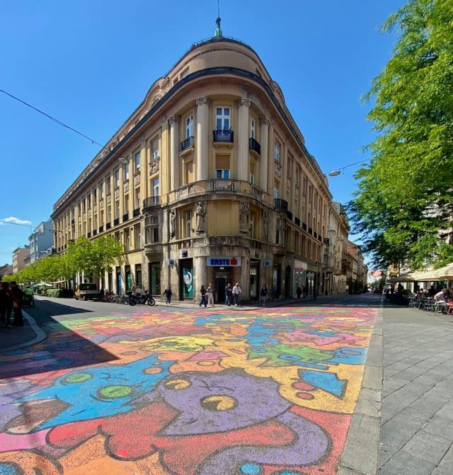 Un mural vibrante de personajes de dibujos animados pintado en un cruce peatonal urbano, frente a un gran edificio esquinero bajo un cielo azul despejado.
