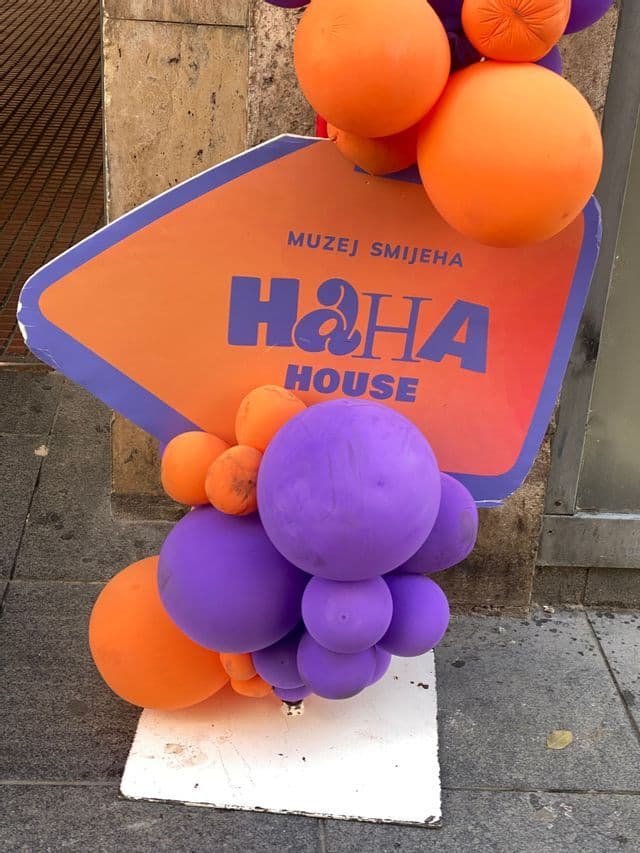 Un cartel de la 'H&HA House, Muzej Smijeha' decorado con globos morados y naranjas en una acera de la ciudad.