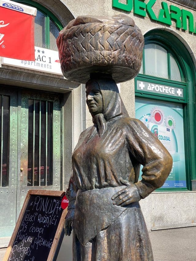 Una estatua de bronce de una mujer con vestimenta tradicional equilibrando una gran cesta tejida en su cabeza en una calle de la ciudad.