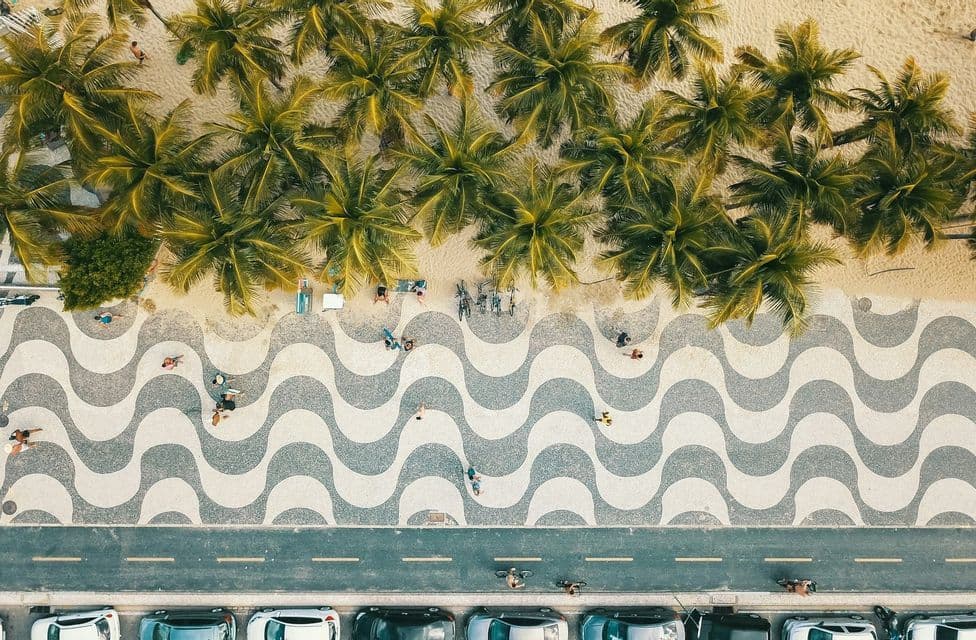 Una vista aerea, dall'alto, di una spiaggia con palme, sabbia e un marciapiede con un caratteristico motivo a onde bianche e nere.