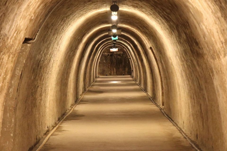 Un túnel subterráneo largo y arqueado con un camino de hormigón, iluminado por una hilera de luces en el techo.
