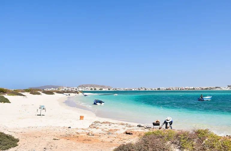 Una persona con un sombrero blanco haciendo una barbacoa en una playa de arena blanca junto a agua turquesa, con un pueblo visible al otro lado de la bahía.