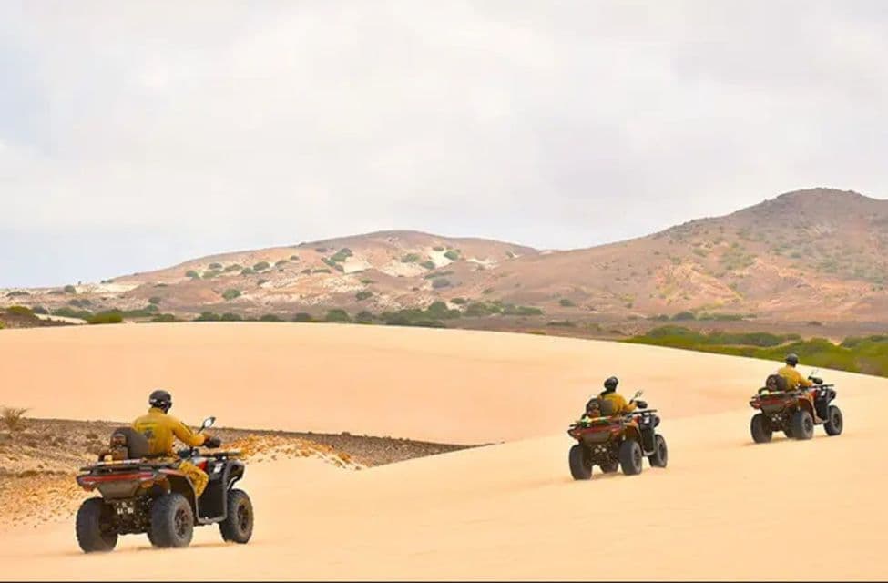 Tres personas de un viaje en grupo de WeRoad conducen cuatrimotos en fila por las dunas de arena del desierto bajo un cielo nublado.
