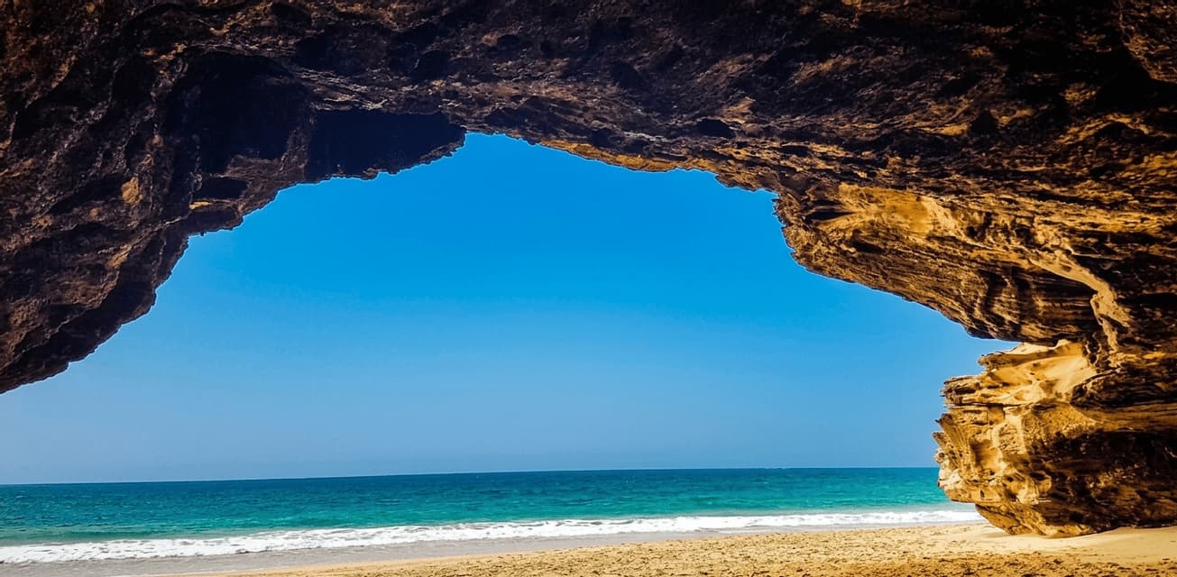 Una vista desde el interior de una cueva de roca se asoma a una playa de arena y un océano turquesa bajo un cielo despejado.
