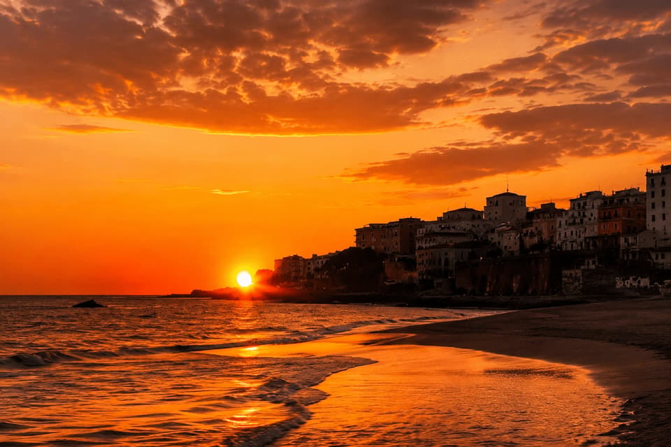 Il sole tramonta sul mare, proiettando un bagliore arancione su una spiaggia sabbiosa accanto a una città collinare sotto un cielo nuvoloso.