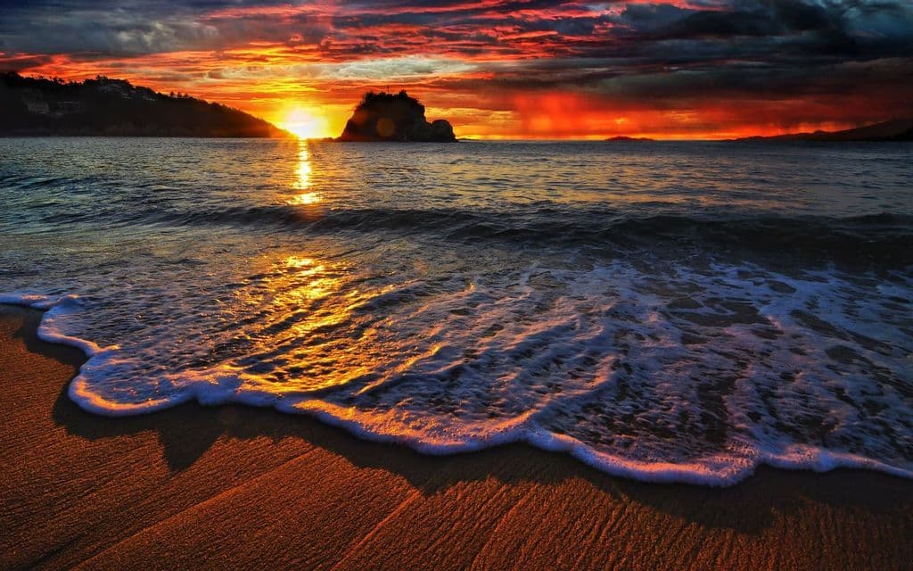 Un'onda si infrange su una spiaggia sabbiosa, illuminata da un tramonto infuocato sull'oceano con colline in silhouette sullo sfondo.