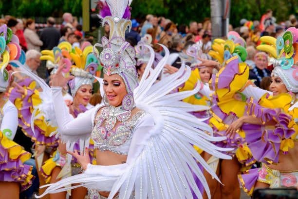 Una donna con un costume di carnevale bianco piumato balla in una sfilata, circondata da altri artisti in abiti colorati.
