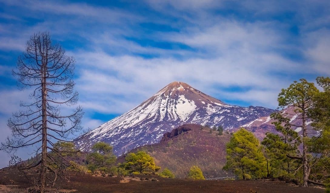 Un vulcano coperto di neve si erge da un paesaggio roccioso punteggiato di pini sotto un cielo azzurro parzialmente nuvoloso.