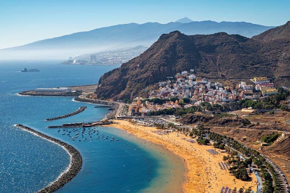 Veduta aerea di una spiaggia dorata e di una cittadina incastonata tra montagne rocciose e l'oceano blu.