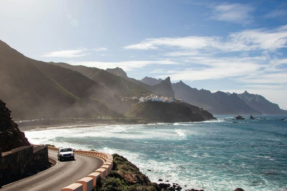 Un'auto percorre una strada tortuosa lungo una costa montuosa, con un piccolo villaggio che si affaccia sull'oceano.