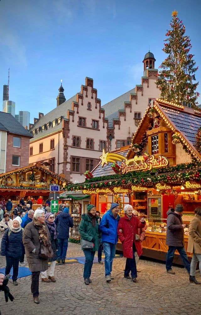 Una folla passeggia in un tradizionale mercatino di Natale, su una piazza acciottolata con bancarelle decorate e un grande albero.